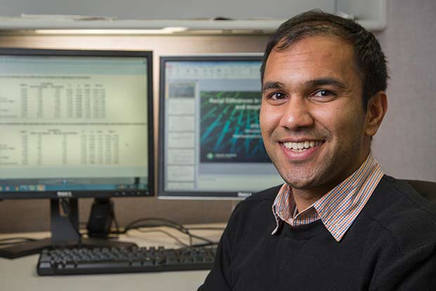 a man, smiling and looking into the camera with computer monitors behind him.